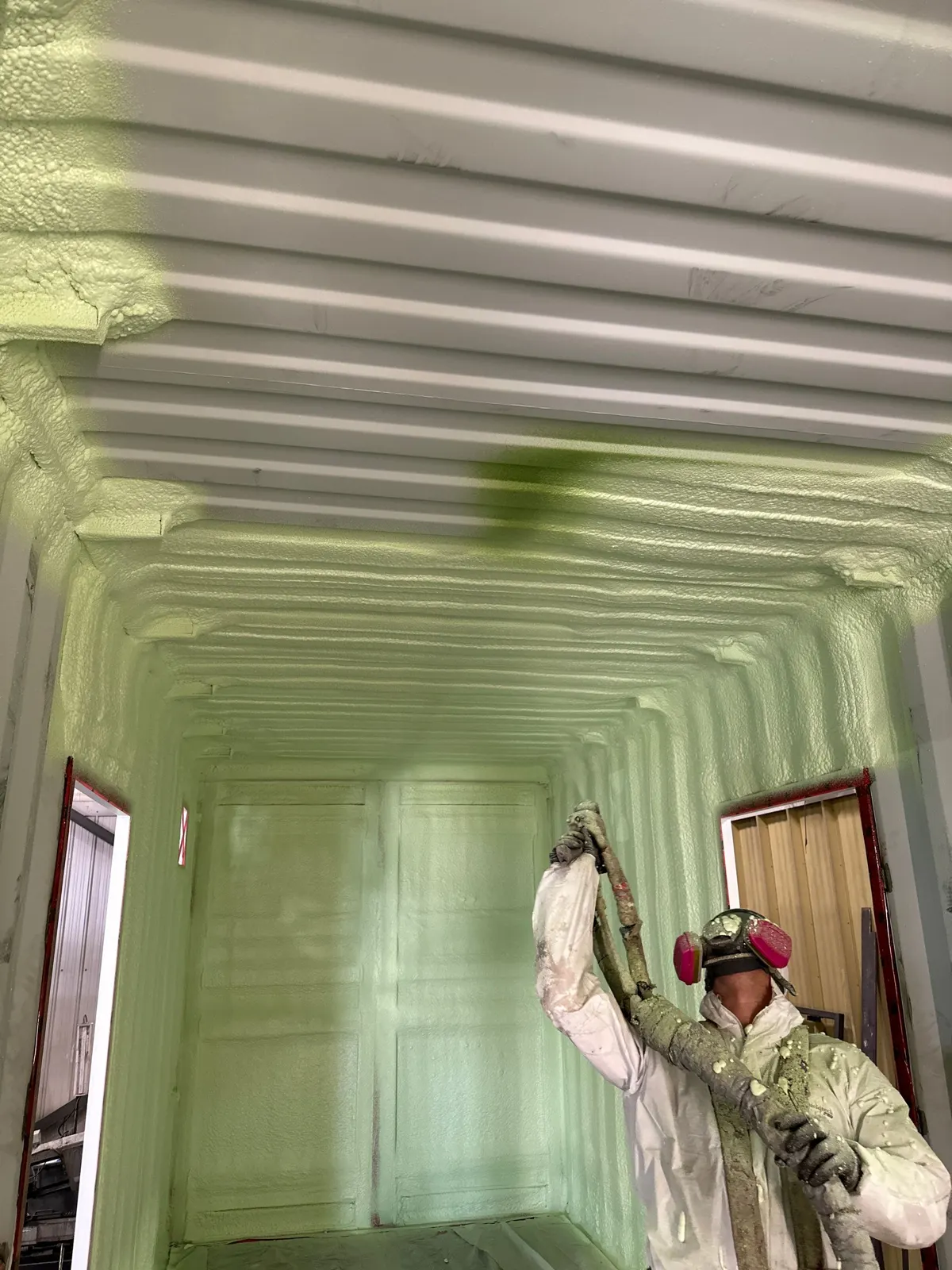 Installer in a suit and respirator spraying green foam inside a shipping container