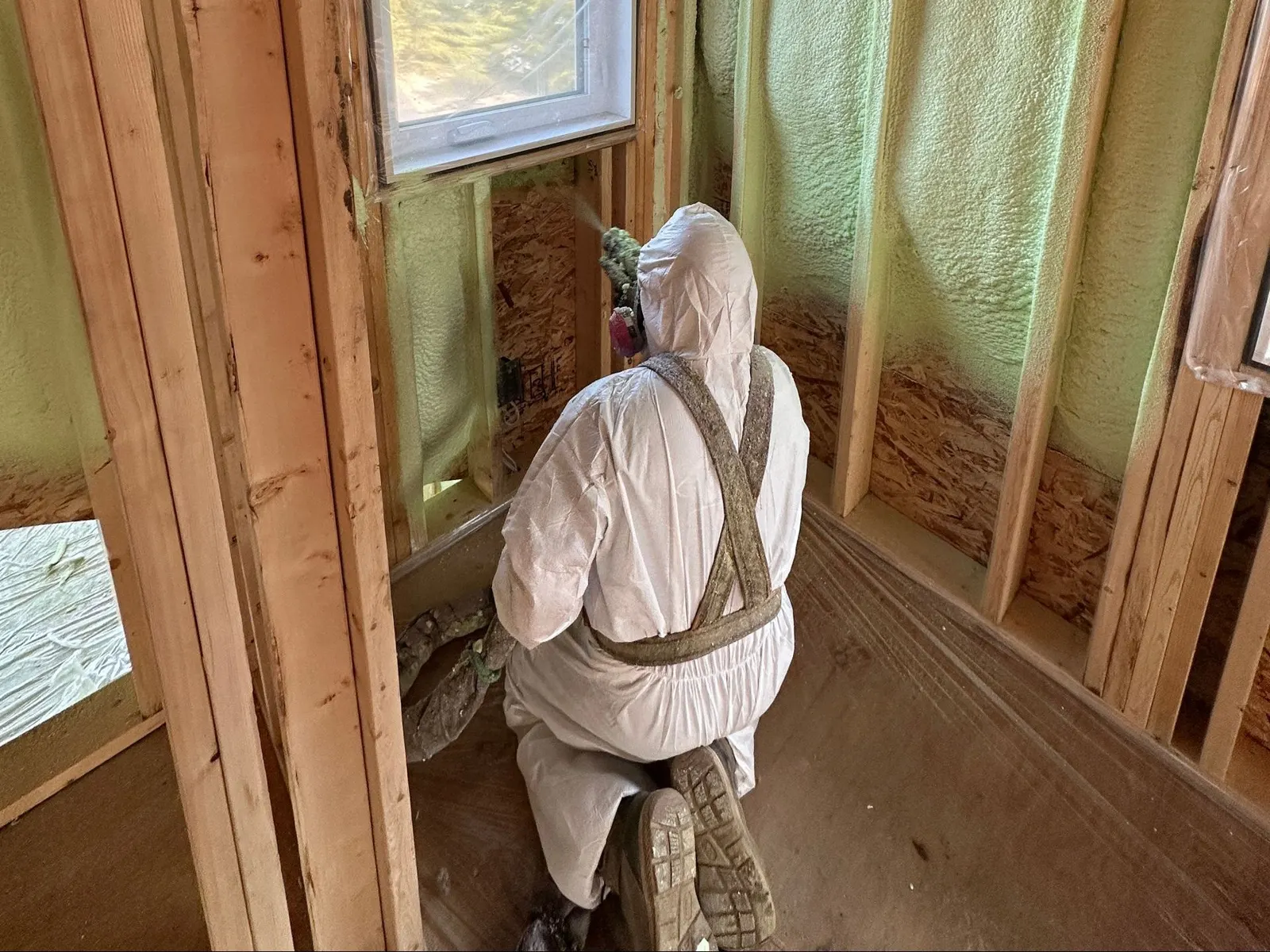 Installer in a protective suit spraying foam into open wall cavities beside a window in a framed room